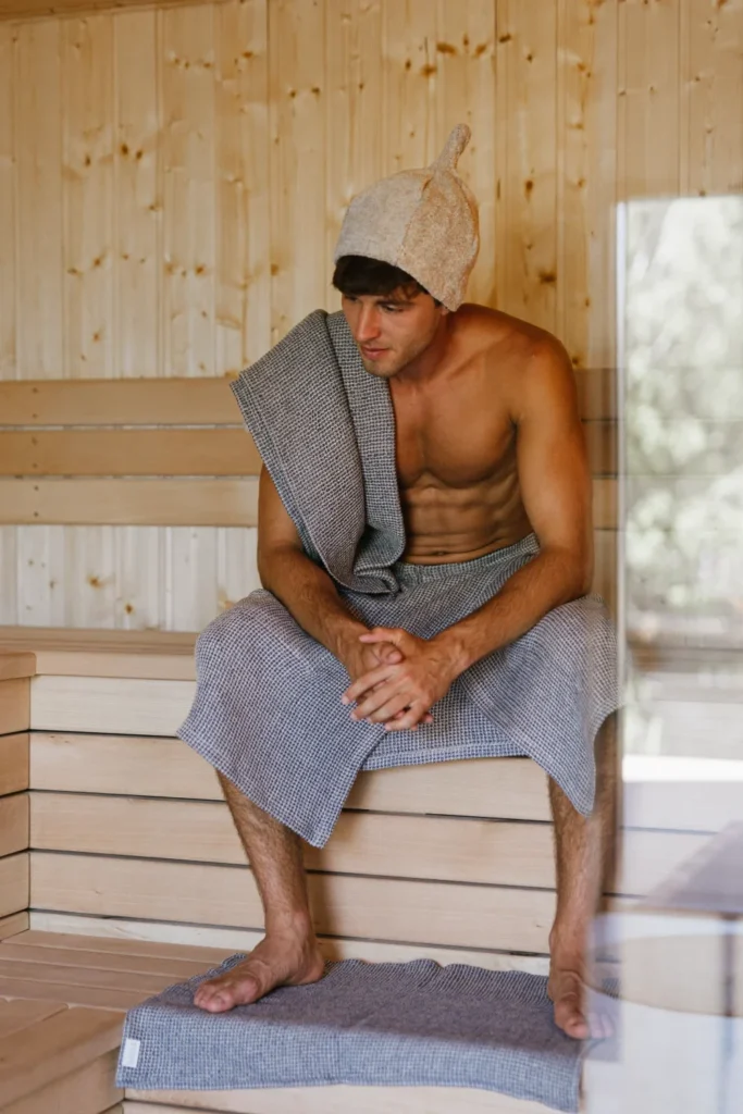 mężczyzna siedzący w saunie ubrany w lniany kilt i owinięty ręcznikiem lnianym wafel natural jeans, w czapce frotte