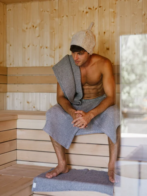 mężczyzna siedzący w saunie, owinięty kiltem do sauny wafel natural jeans blue i ręcznikiem do sauny, ubrany w czapkę do sauny
