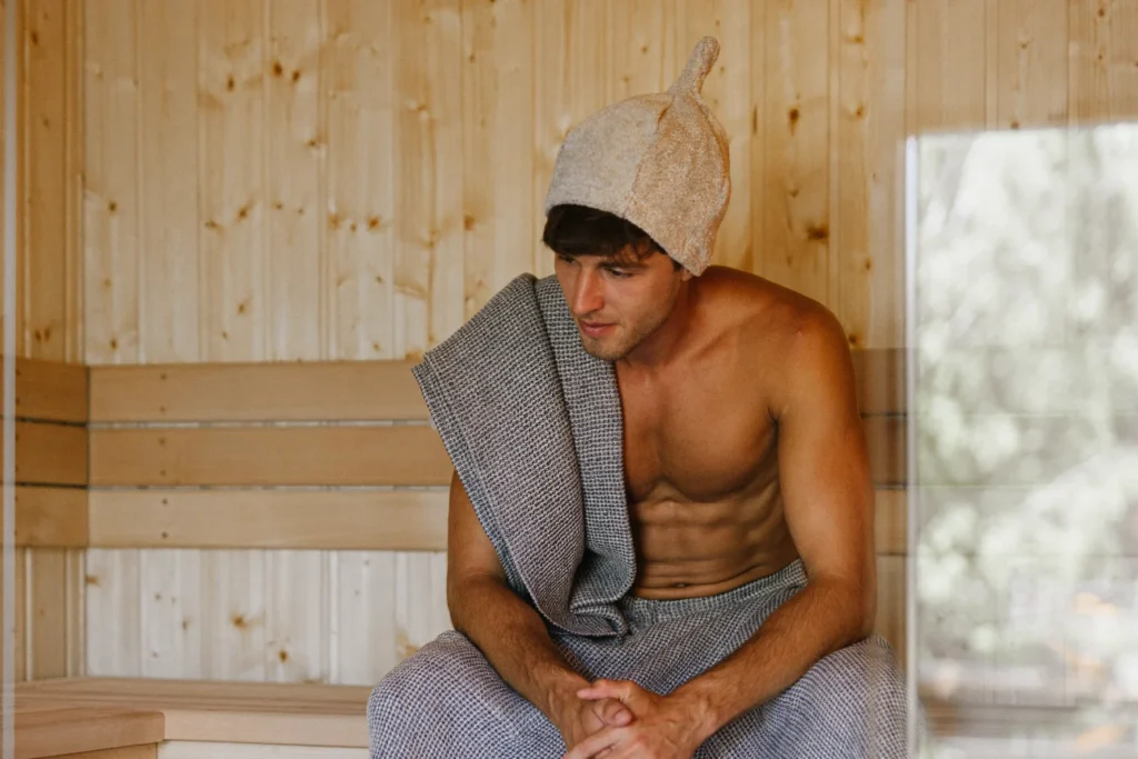 Mężczyzna w trakcie saunowania z czapką do sauny frotte natural, owinięty ręcznikiem i ubrany w kilt do sauny